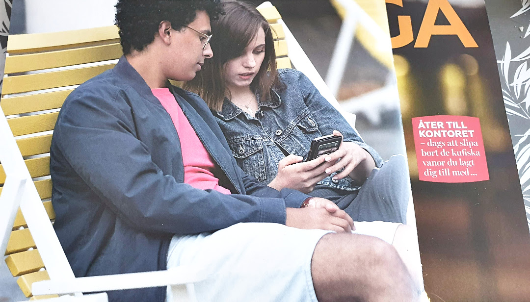 Casal de jovens sentados em uma cadeira grande olhando juntos a tela de um telefone.  Ele de jaqueta azul, camiseta rosa e bermuda cinza.  Ela de jaqueta jeans azul.