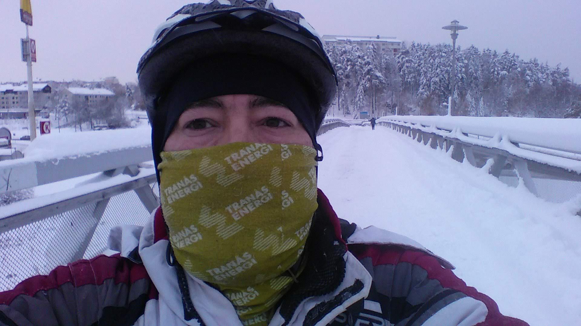 Foto minha com roupas e capacete que uso quando ando de bicicleta sobre uma ponte coberta de neve.
